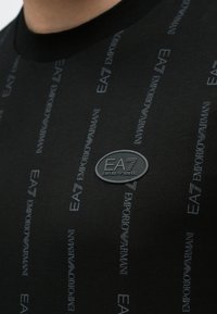 Fekete ing világosszürke EA7 Emporio Armani logókkal ismételve, középen a mellkashoz közel egy ovális EA7 Emporio Armani jelvénnyel.