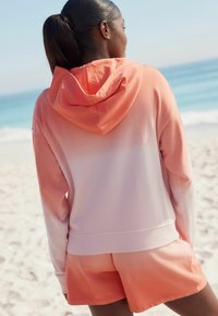 Conjunto de sudadera y pantalones cortos en colores coral y melocotón degradados. Tejido suave, corte relajado con capucha. Textura suave; mínimo detalle.