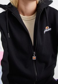 Personne portant un sweat à capuche noir zippé avec des accents blancs et roses sur les manches et un logo ellesse sur la poitrine et la tirette de la fermeture éclair.
