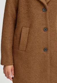 Bruine wollen jas met een getextureerd oppervlak, zwarte knopen en een appliekzak aan de voorkant. Het ontwerp bevat een kraag en lange mouwen.