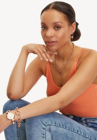 Schmuck aus Roségold umfasst eine klobige Kettenhalskette, Creolen und eine Uhr mit rundem Ziffernblatt. Das Oberteil besteht aus orangefarbener gerippter Stoff.