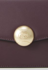 Bordeaux læderpung med en guld-tone rund lukning med prægede Lacoste-logo og kontrasterende syninger langs kanterne.