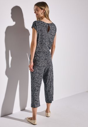 Marineblauer Jumpsuit mit weißem Wellendesign, kurzen Ärmeln, Schlüsselbereich am Rücken und verkürzten Beinen. Kombiniert mit beigen Ballerinas. Glatte Stoffoberfläche.