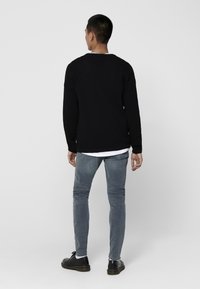 Maglione nero lavorato a maglia con maniche lunghe, abbinato a jeans skinny blu chiaro e scarpe nere stringate. Lo sfondo è di un bianco uniforme.
