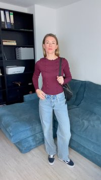 Bordeauxfarbene Langarmbluse mit strukturierten Details, kombiniert mit hellblauen weit geschnittenen Jeans und schwarzen Sneakern, stehend in einem Raum mit einer Couch.