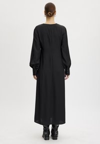 Largo vestido negro hecho de una tela suave, con mangas largas abullonadas y acentos de botones en los puños. Escote redondo sencillo, con detalles mínimos.
