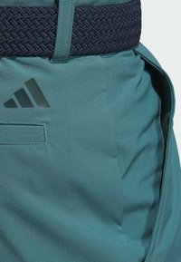 Teal shorts with a woven navy waistband, featuring an adhesive black logo and a side pocket. Smooth texture, tailored fit.