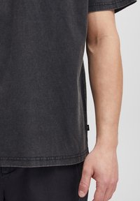 Tricou negru cu mânecă scurtă, fabricat dintr-un material moale, având un croi lejer, cusături vizibile și o etichetă pe lateral.