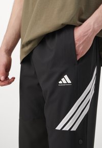 Person wearing black basketball shorts with three white stripes and a logo, hand resting inside right pocket, paired with olive green shirt.