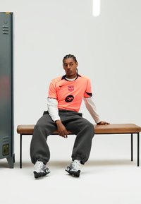 Ryškiai rožinė sportinė liemenė virš balto ilgomis rankovėmis marškinėlių, kartu su pilkomis sportinėmis kelnėmis ir juodo-baltos spalvos sportbačiais, sėdint ant medinės suolelio.