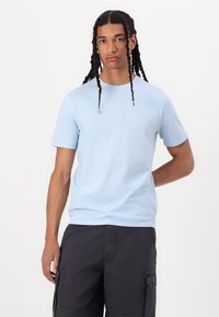 Μπλε βαμβακερό t-shirt με στρογγυλή λαιμόκοψη και κοντά μανίκια, εκτεθειμένο σε μοντέλο που φορά μαύρες cargo βερμούδες, σε ένα απλό φόντο.