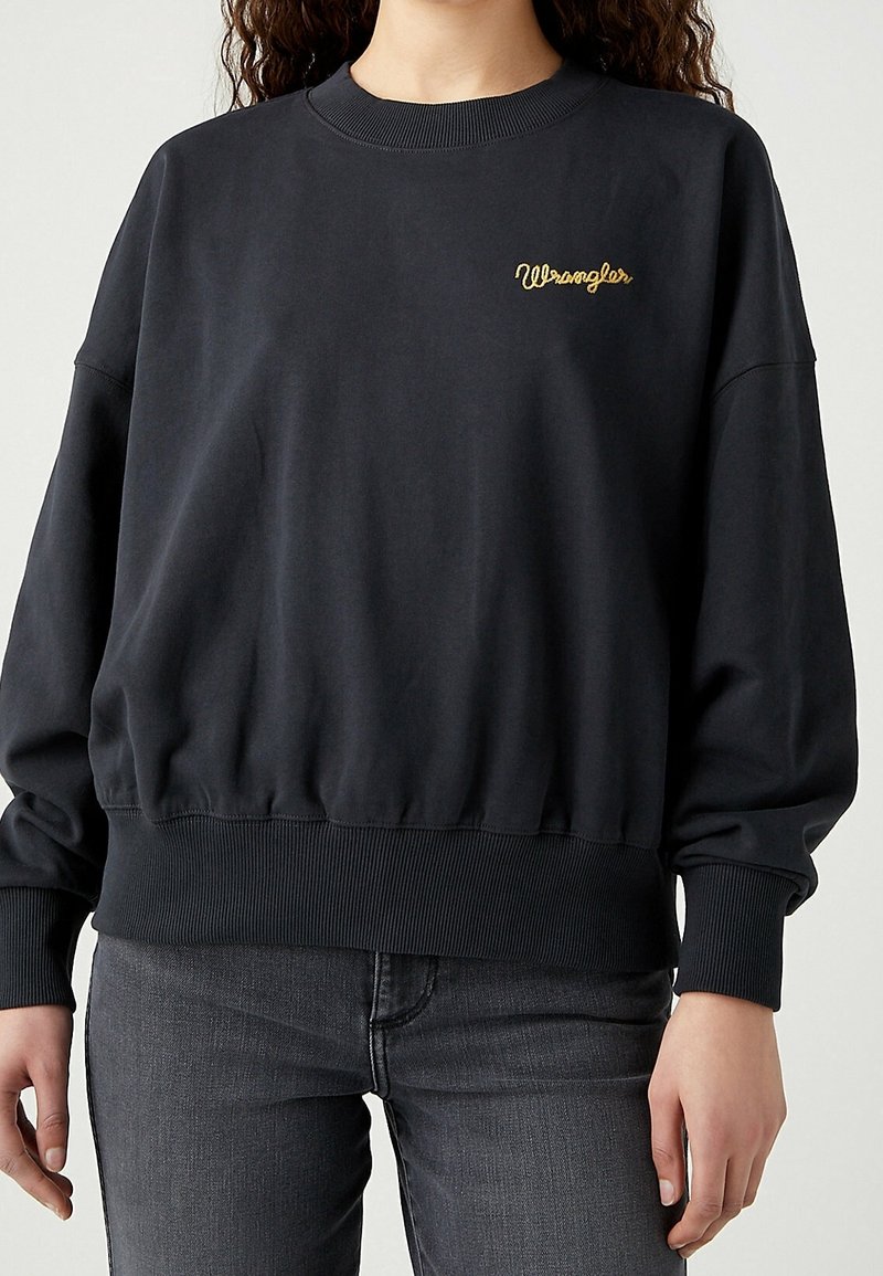 Femme portant un sweat-shirt noir Wrangler avec un logo jaune, associé à un jean gris foncé, debout devant un fond uni.