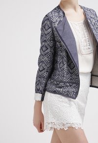 Veste à motif marine avec un devant zippé, tissu texturé, col gris et poignets repliés, associée à une jupe en dentelle blanche avec des bords festonnés.