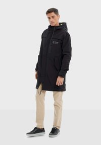 Parka noire avec capuche, dotée d'une fermeture éclair, de poches avant et d'une étiquette. Portée sur un pantalon beige et des baskets grises.