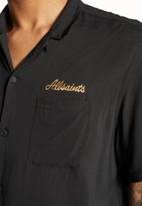 Svart kortärmad skjorta i slätt tyg med en ficka och broderad "Allsaints"-logotyp i guld på bröstet.