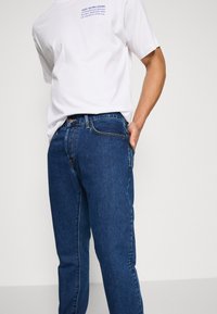 Hvit t-skjorte med blå tekst, kombinert med mørkeblå denimjeans. Jeansene har rett passform og et klassisk femlommersdesign.