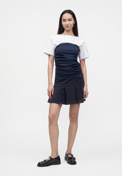 Femme debout devant un fond uni, portant un top blanc à manches courtes sous une robe bleu marine froncée et une jupe plissée bleu marine avec des mocassins noirs.