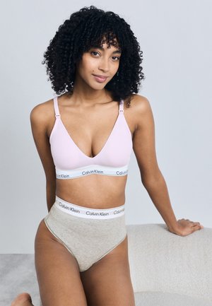 Kvinde med krøllet hår iført en lyserød bh og grå trusser med høj talje, begge med Calvin Klein-branded taljebånd, lænende sig op ad møbler.