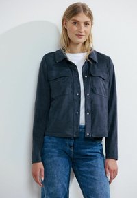 Giacca in suede blu navy con colletto, chiusura frontale con bottoni a pressione e due tasche sul petto; abbinata a jeans in denim blu chiaro.