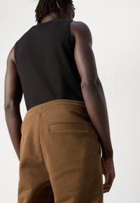 Person wearing a black sleeveless top and brown pants with a back pocket, shown from behind against a plain light background.