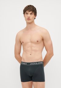 Črne bokserice z Jacquard pasom, na katerem je napis "JACK & JONES." Material se zdi mehak, oblikovana zasnova z ravnimi šivi in prilegajočo konturo.