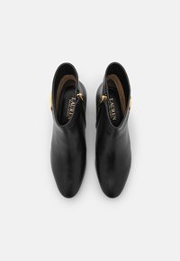 Un par de botas de cuero negras hasta el tobillo con cremalleras doradas en los lados, punta redondeada y plantillas "Lauren Ralph Lauren" vistas desde arriba sobre un fondo blanco.