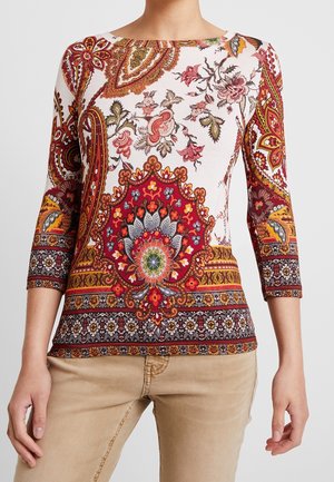 Frau trägt ein weißes, rotes und mehrfarbiges Top mit Paisley- und Blumenmuster, dreiviertellangen Ärmeln, kombiniert mit beigen Hosen.