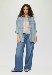 Hellblaue Jeansjacke über einem grauen Shirt mit Sonnenstrahlgrafik, kombiniert mit weiten blauen Jeans und grauen Schuhen, stehend vor einem neutralen Hintergrund.
