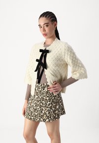 Cardigan in maglia crema con un motivo testurizzato, chiusura con fiocco in velluto nero, indossato sopra un top marrone a coste e una mini gonna con stampa leopardata.