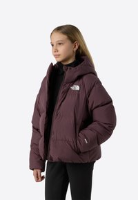 Vijolično puhovka s kapuco, opremljena z zadrgo spredaj, elastiki na manšetah in belo nalepko na levi prsni strani. Gladek, prešit teksturiran material.