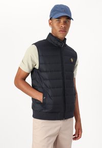 Black puffer vest with high collar, zip closure, side pockets, and logo patch; worn over a light beige short-sleeve shirt. Additional blue cap.