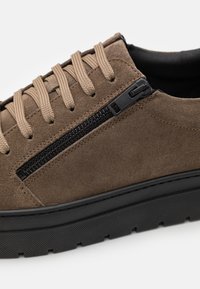 Baskets en daim marron avec des lacets beiges et un détail de fermeture éclair noire. Elles présentent une semelle en caoutchouc noire texturée, offrant durabilité et adhérence.