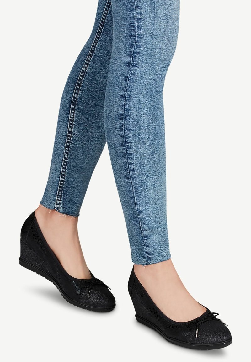 Jambes en jean bleu ajusté portant des ballerines compensées noires avec petits nœuds et finition texturée sur fond neutre.