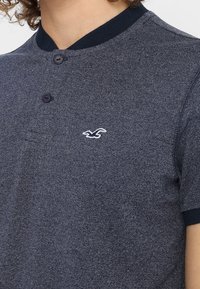 Marinblå polotröja med kort ärm i textilerad fabric, tvåknappsknäppning och en vit broderad logotyp på bröstet. Kontrastkrage.