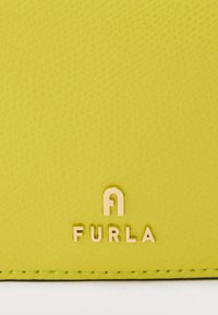 Gros plan sur un cuir texturé jaune vif avec le logo doré Furla et des coutures le long du bord inférieur.