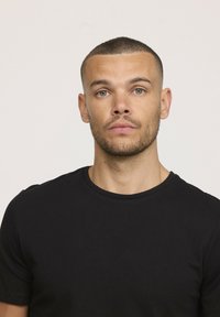 T-shirt noir à manches courtes en coton, avec un col rond. Portrait d'une personne avec des cheveux courts et une barbe, sur fond neutre.