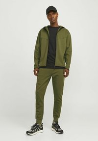 Olive green athletic tracksuit with a zip-up hooded jacket, slim-fit pants, and black accents, paired with black sneakers. Smooth fabric texture.