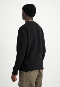 Felpa nera a maniche lunghe, con polsini a coste e vestibilità rilassata. Indossata con pantaloni cargo verde oliva. Berretto in testa. Sfondo bianco.