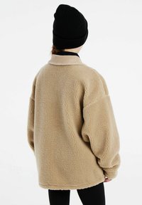 Pull en polaire beige avec une surface texturée, coupe oversized et un col en velours côtelé de couleur claire. Porté avec un legging noir et un bonnet noir.