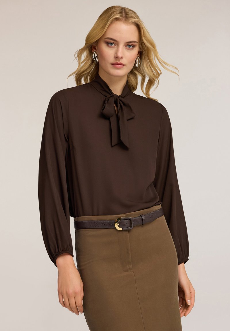 Blusa marrón de manga larga con cuello de lazo, combinada con una falda lápiz marrón y un cinturón oscuro con hebilla dorada.