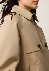 Beige trench coat, jossa on napitettavat etuosat, leveä kaulus ja sileä rakenne. Varustettu mustilla napeilla ja sivutaskuilla.