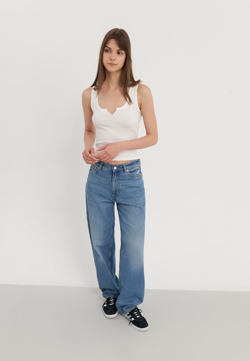 Top in cotone bianco a costine con scollatura ondulata, abbinato a jeans a gamba larga blu chiaro. La modella indossa sneakers nere. Tessuto testurizzato.