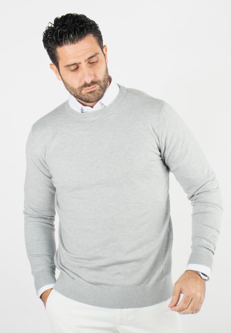 Un pull léger gris clair avec un col rond et des manches longues, présentant un ourlet et des poignets côtelés. Porté par-dessus une chemise blanche à col.
