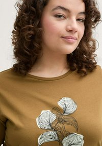 Bruine T-shirt met korte mouwen en een ronde halslijn, voorzien van een zwart lijntekeningen bloemenontwerp aan de voorkant, gemaakt van een zachte, gladde stof.
