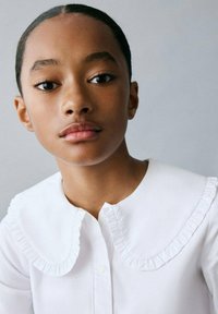 Cămașă albă cu guler volant, având închidere cu nasturi. Textură netedă a materialului pe un fundal neutru.