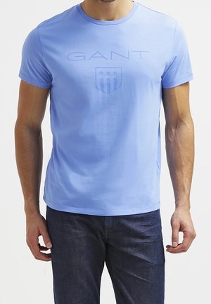 Man wearing light blue GANT short-sleeve t-shirt and dark blue jeans, standing against plain background.
