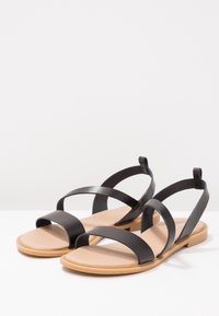 Sandales plates noires avec plusieurs brides, semelle de couleur beige, et un design minimaliste. Comprend un petit talon et une semelle texturée.