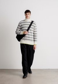 Strikkegenser med geometriske mønstre i svart og hvitt, høyhalset, lysegrønne detaljer, kombinert med løstsittende sorte bukser og en skulderveske.