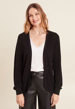 Cardigan noir en tricot avec un devant ouvert, manches longues et poignets côtelés. Porté par-dessus un top blanc avec dentelle et un pantalon en cuir noir.