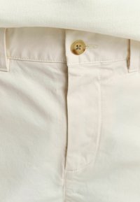 Lichtbeige broeken met een zichtbare knoopsluiting, gestikte details en een gladde textuur. De stof lijkt duurzaam en zacht.